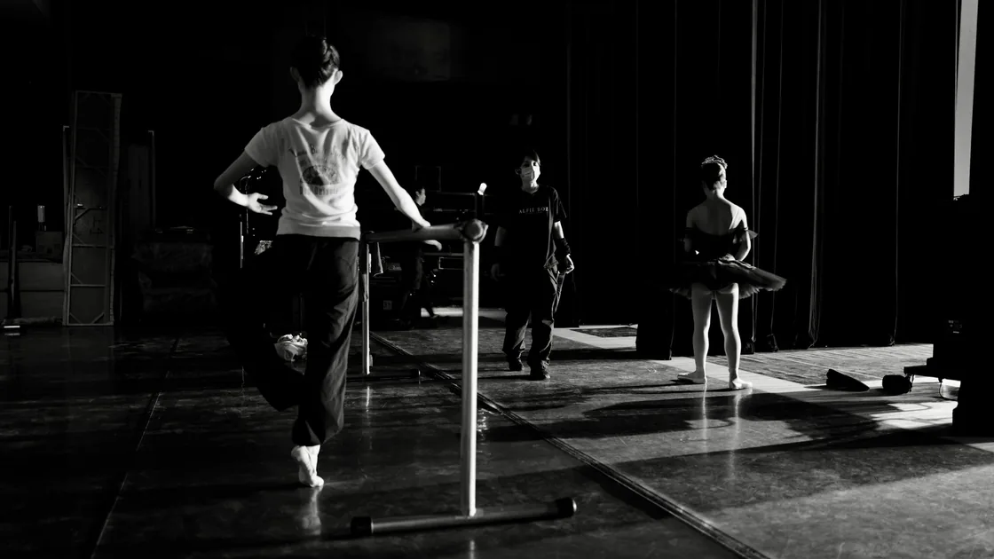 Sala de ensayo en penumbra con bailarines trabajando junto a una barra